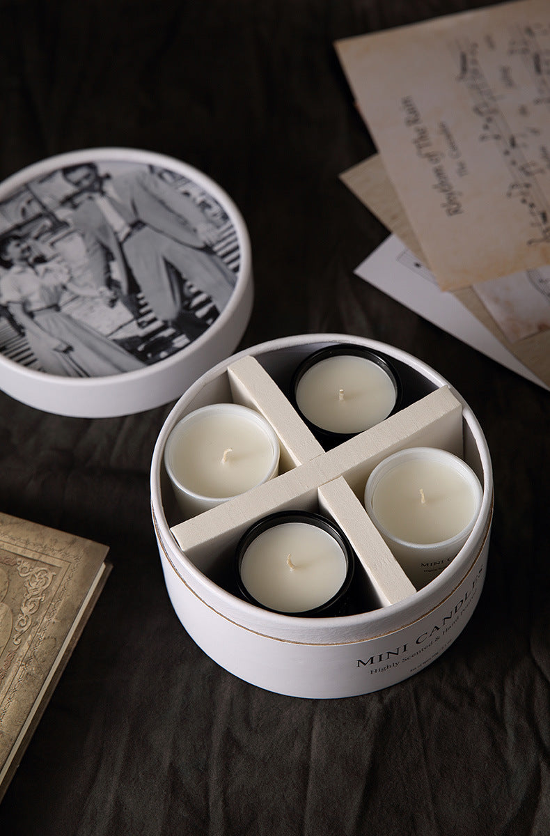 Round white container with four lit candles on a dark surface with vintage books and papers.