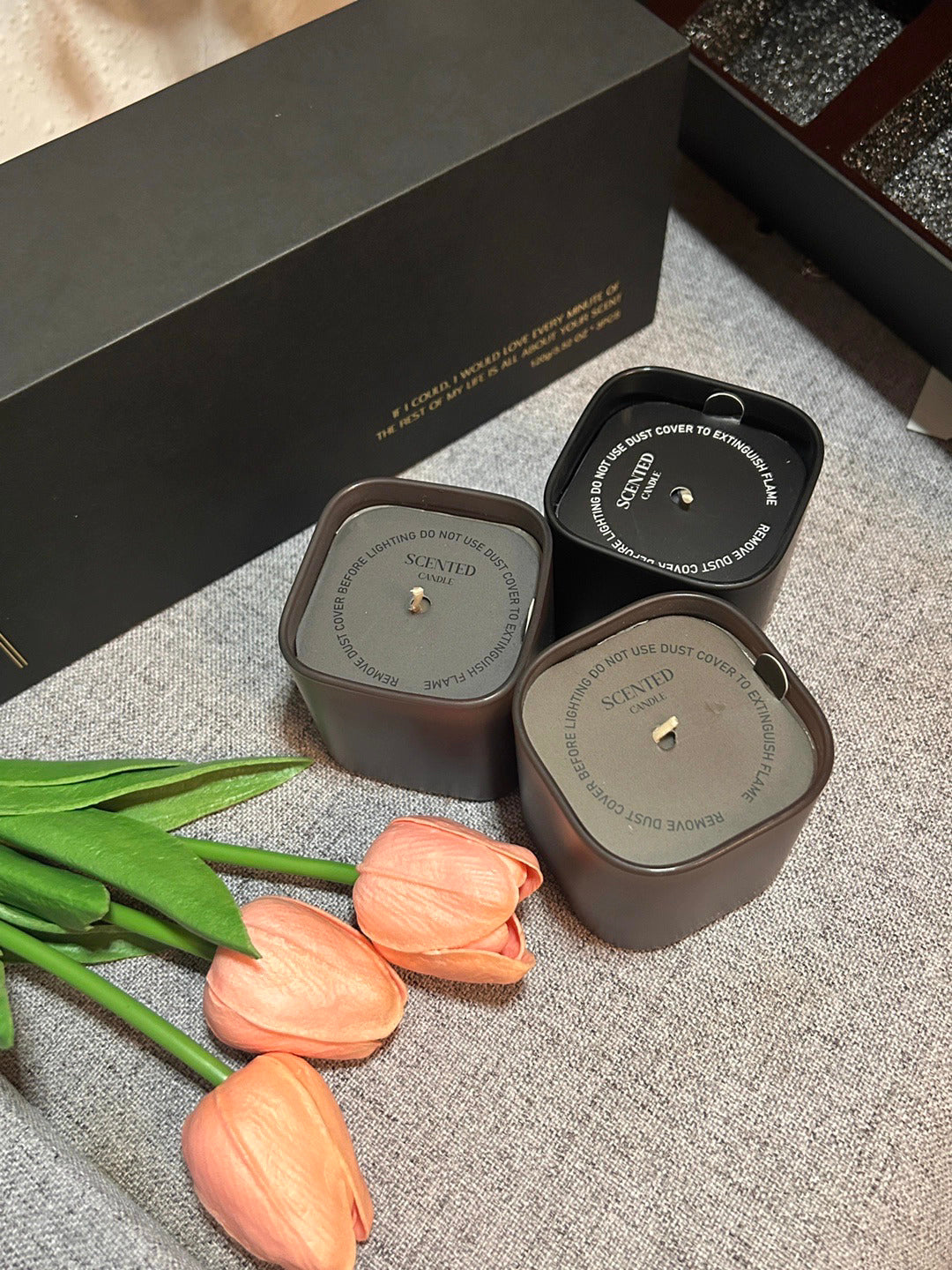 Two scented candles in black containers with pink tulips on a textured surface