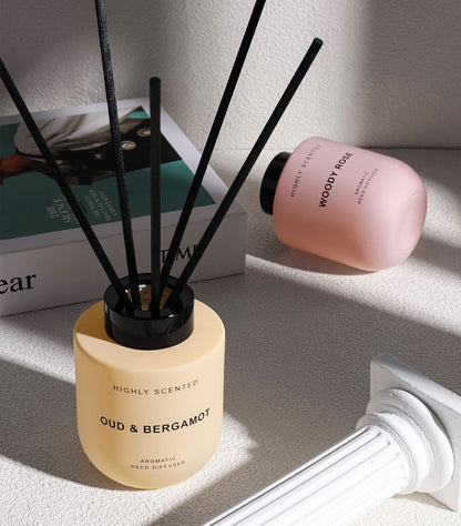 Two diffusers, one beige with black sticks labeled 'Oud & Bergamot' and the other pink with black text labeled 'Woody Rose', on a light surface.