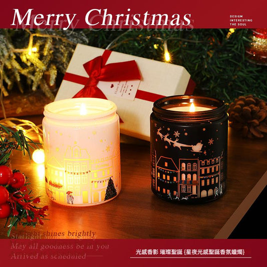 Two lit candles with Christmas designs on a wooden surface, surrounded by festive decorations.