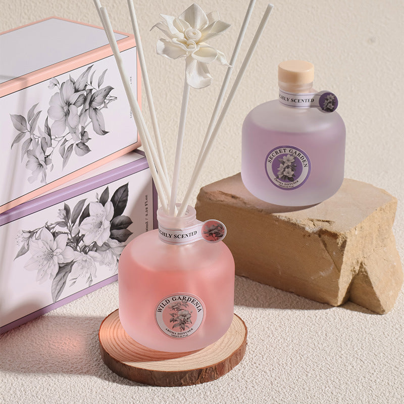 Pink diffuser with reeds and a bottle on a wooden block, with a floral-themed book in the background.