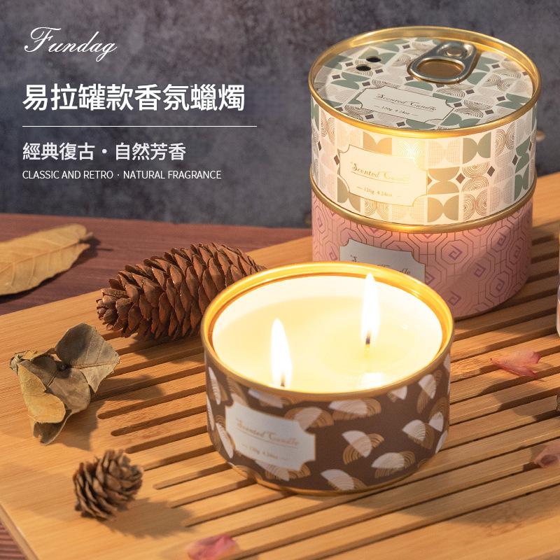 Candle in a decorative tin with a pattern, surrounded by pine cones and leaves on a wooden surface.
