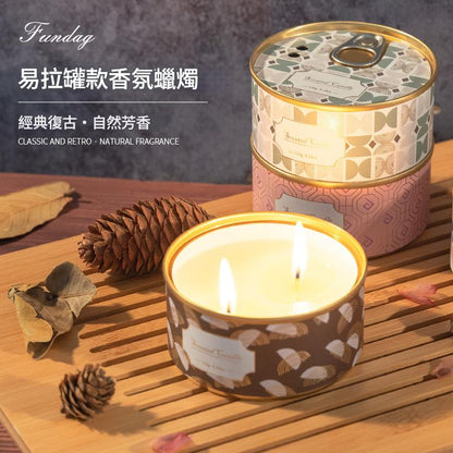 Candle in a decorative tin with a pattern, surrounded by pine cones and leaves on a wooden surface.