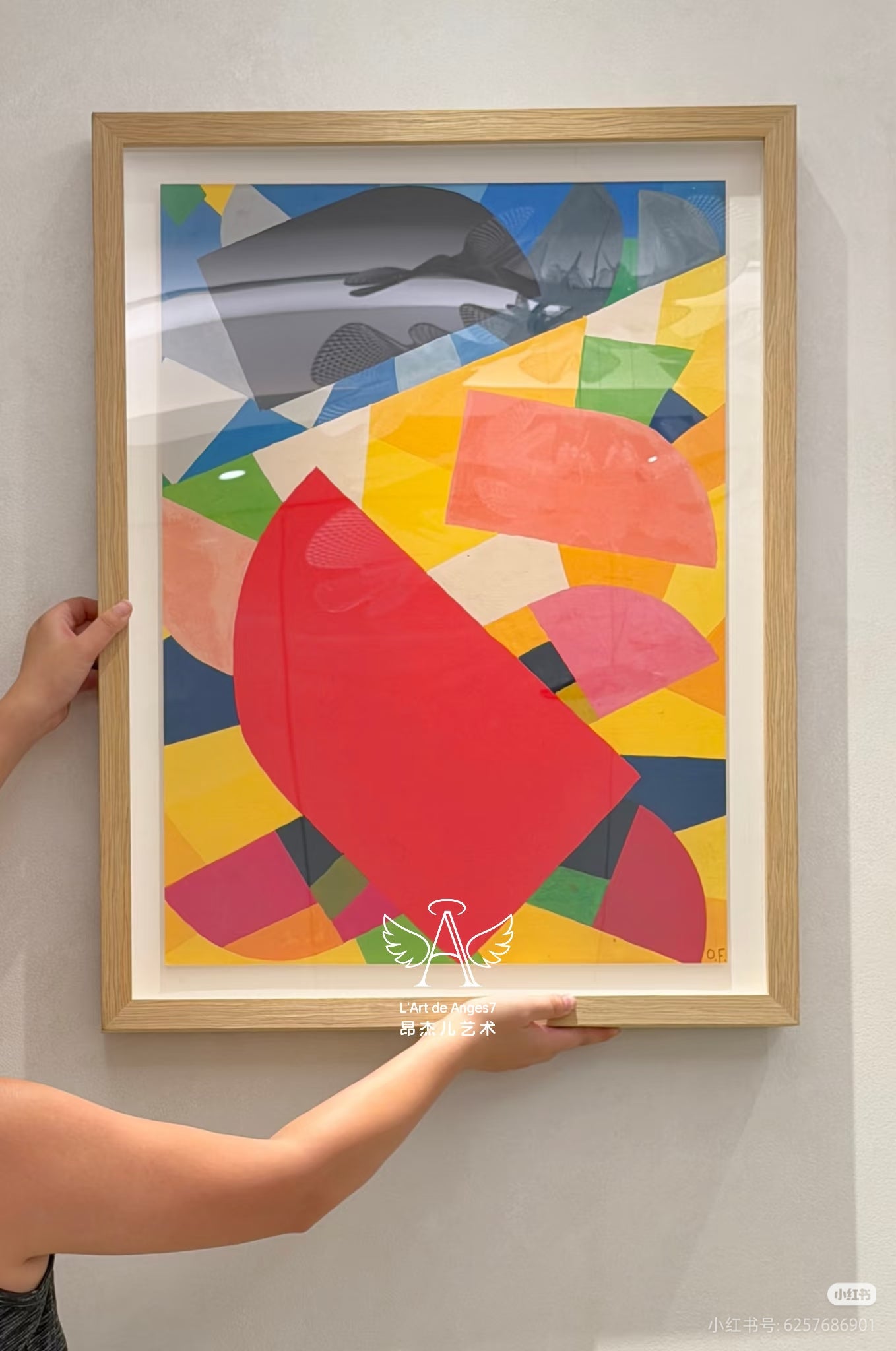 Person holding a colorful abstract art print in a wooden frame against a white wall.