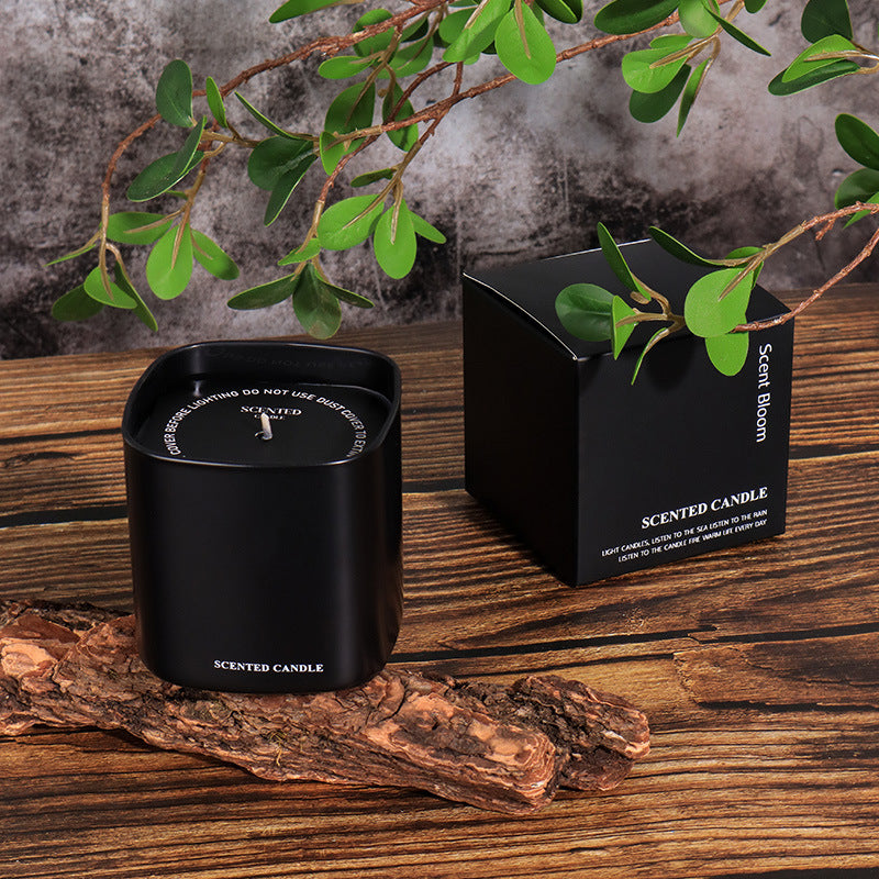 Black scented candle and packaging on a wooden surface with green leaves.