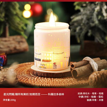 Decorative candle with a building design on a wooden coaster, surrounded by cinnamon sticks and blurred Christmas decorations.