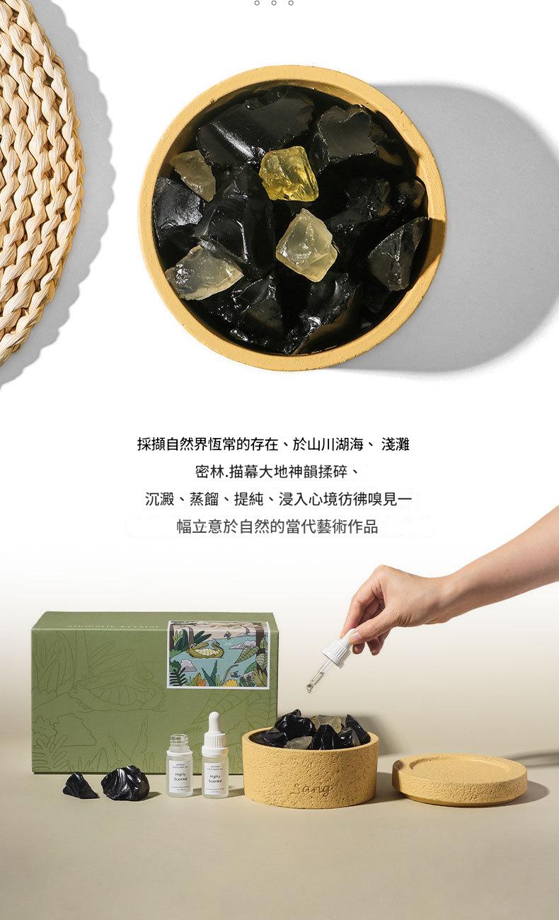 Wooden bowl with dark-colored stones, a green box, and a hand holding a dropper bottle on a light background.
