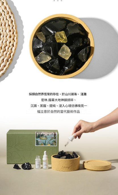 Wooden bowl with dark-colored stones, a green box, and a hand holding a dropper bottle on a light background.