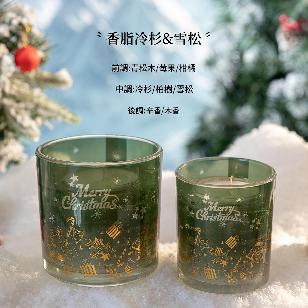 Two green glass candles with 'Merry Christmas' text on a snowy surface with blurred Christmas trees in the background.