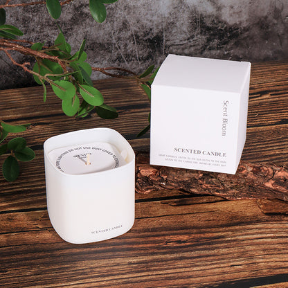 White scented candle and its packaging on a wooden surface with green leaves.