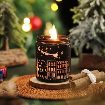 Decorative candle with house and sleigh design on a woven mat with Christmas trees in the background.